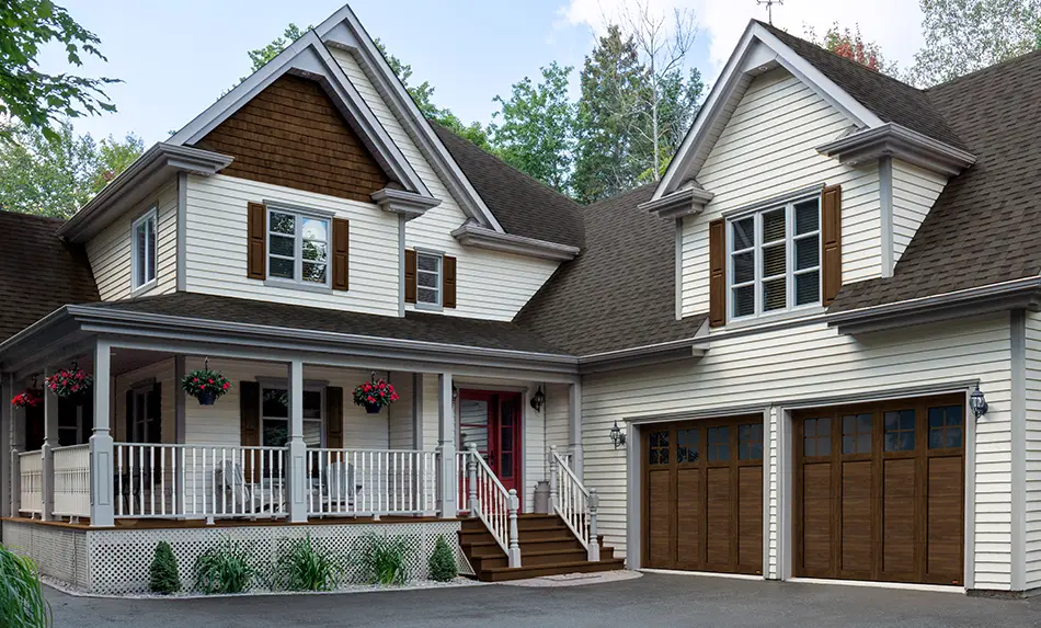 A traditional farmhouse with Walnut shutters and porch is enhanced by Garaga steel garage doors and PVC overlays in Faux-Wood Chocolate Walnut color.